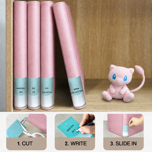 Pink toploader binders on a shelf with cut, write, and slide-in spine labels beside a Mew plush