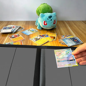 Hand placing a holographic Pokemon card into a binder on a wood table beside a Bulbasaur plush and scattered cards