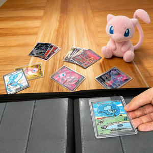 Hand loading a toploadered Pokemon card into a pink toploader binder beside a Mew plush on a wood table