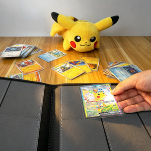 Hand placing a toploadered Pokemon card into yellow 9-pocket binder beside Pikachu cards on wood table