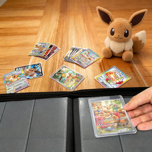 Hand loading a toploadered Pokemon card into a pink toploader binder beside an Eevee plush on a wood table