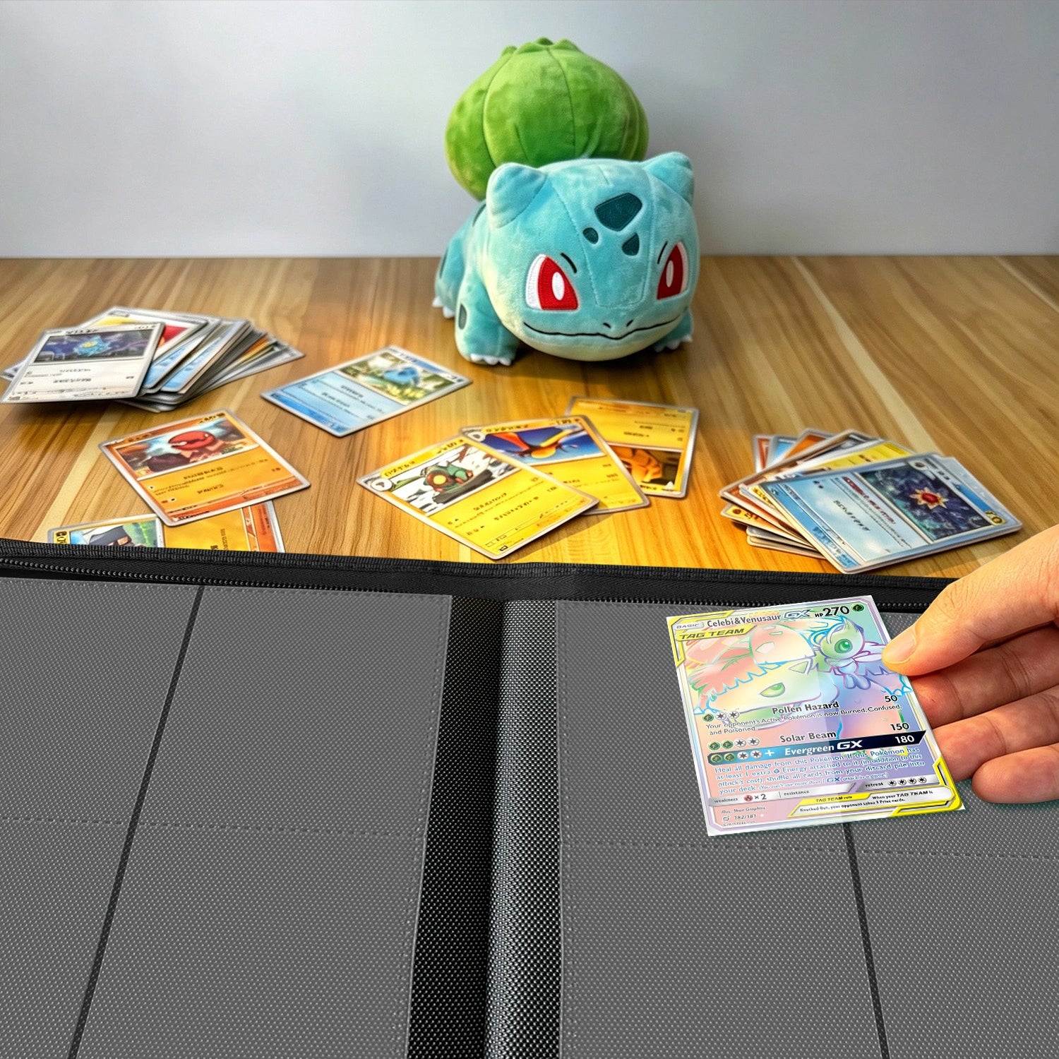 Hand placing a holographic Pokemon card into a binder on a wood table beside a Bulbasaur plush and scattered cards