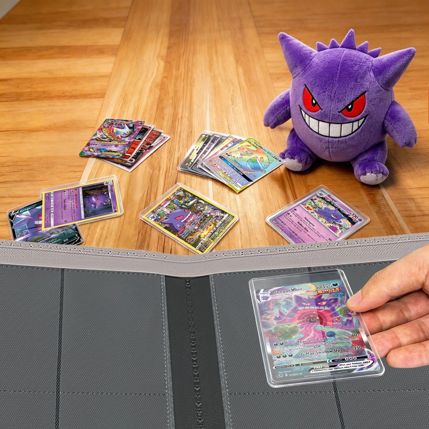Hand placing a toploadered Pokemon card into a binder on a wood table beside a Gengar plush and scattered cards