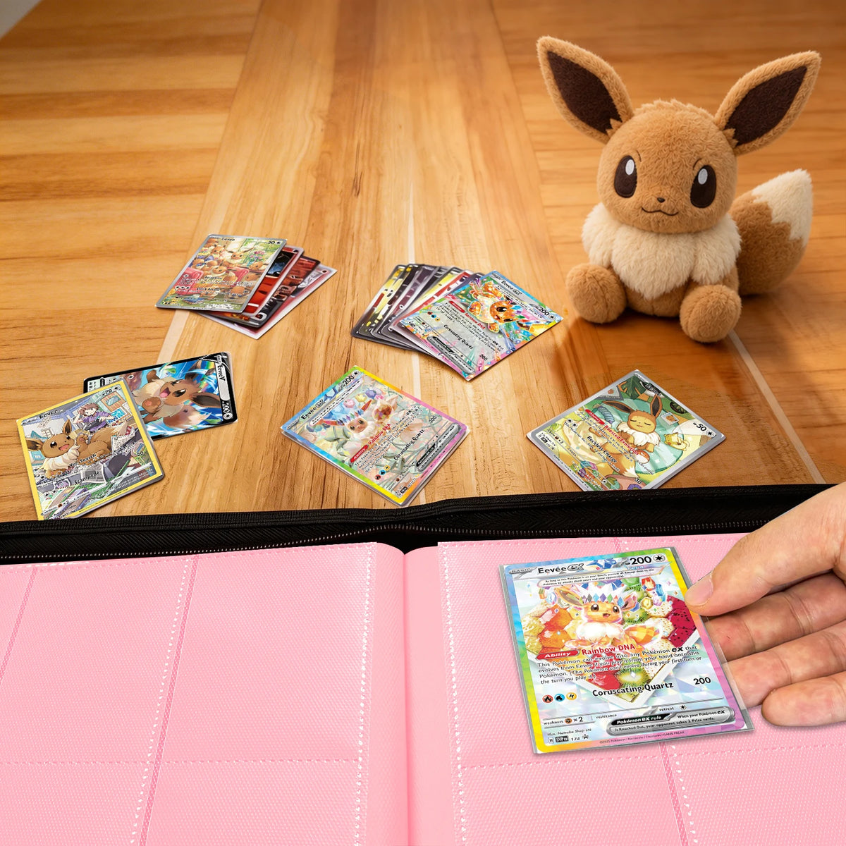 Hand placing a Pokemon card into a pink Eevee Family card binder beside an Eevee plush and scattered cards on a wood table