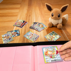 Hand placing a Pokemon card into a pink Eevee Family card binder beside an Eevee plush and scattered cards on a wood table