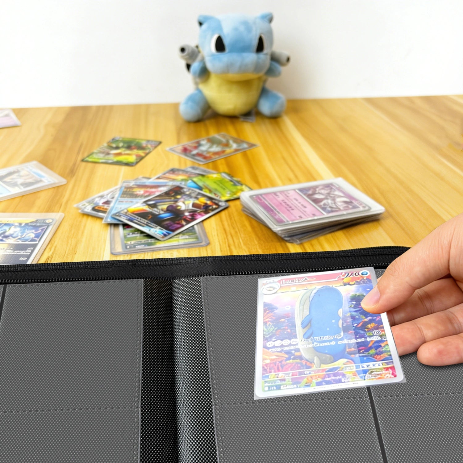 Hand placing a holographic Pokemon card into a binder on a wood table beside a Squirtle plush and scattered cards