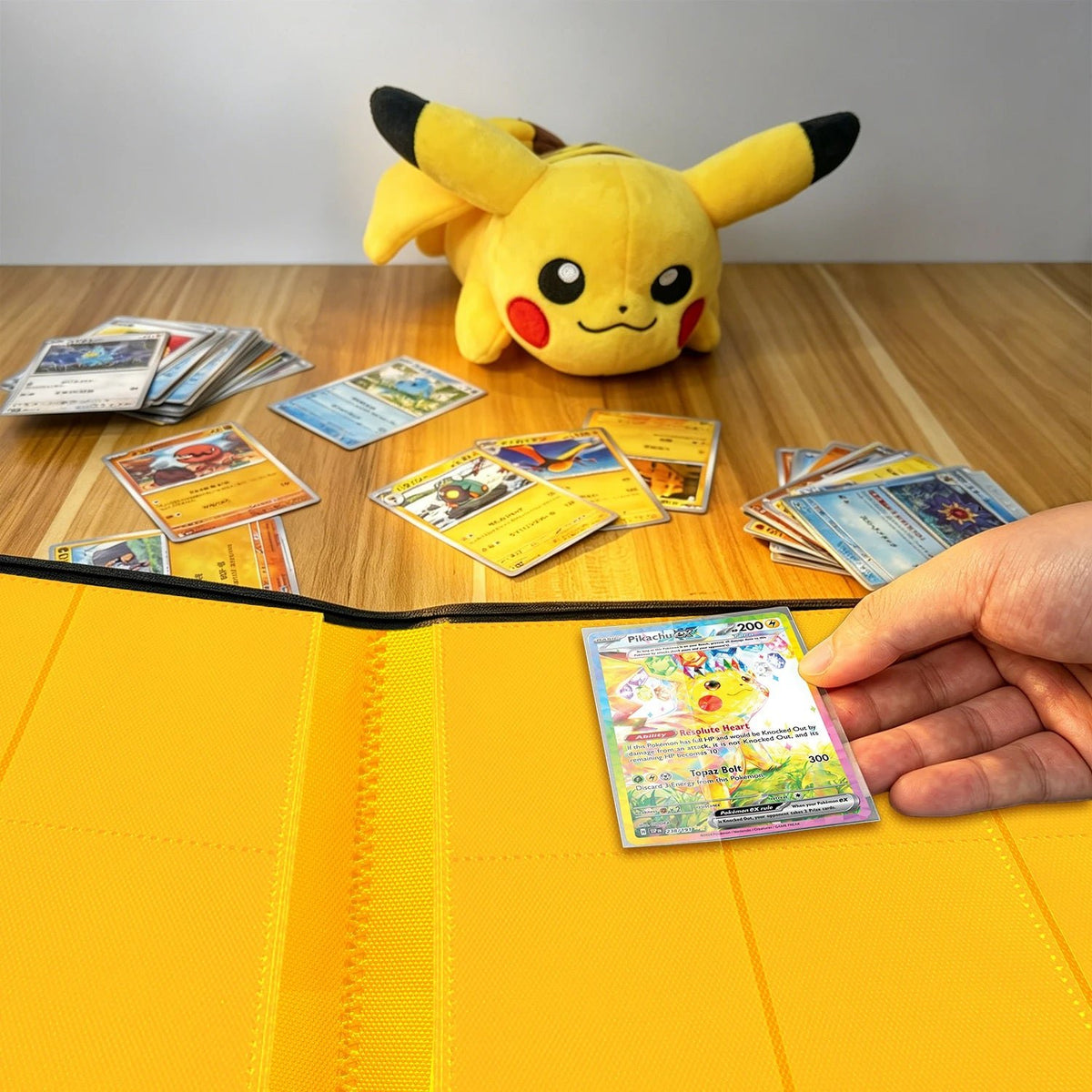 Hand placing a Pokemon card into a yellow card binder beside a Pikachu plush and scattered cards on a wood table