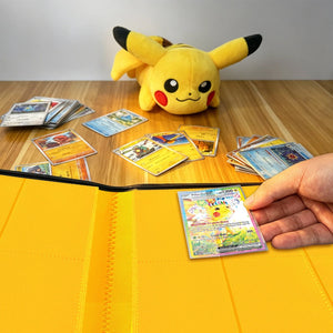 Hand placing a Pokemon card into a yellow card binder beside a Pikachu plush and scattered cards on a wood table