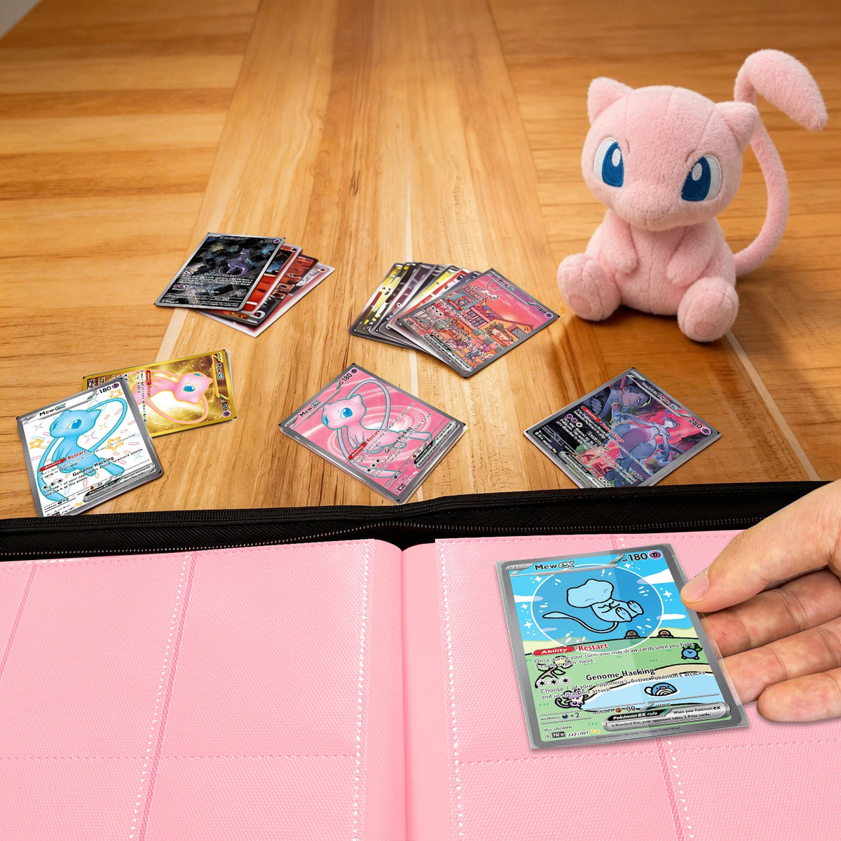 Hand placing a Pokemon card into a pink Mew card binder beside a Mew plush and scattered cards on a wood table