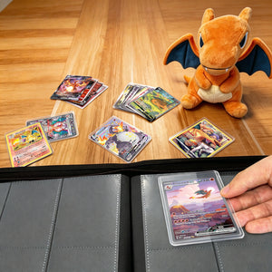 Hand placing graded Pokemon card in toploader into binder beside scattered Charizard cards and plush on wooden table