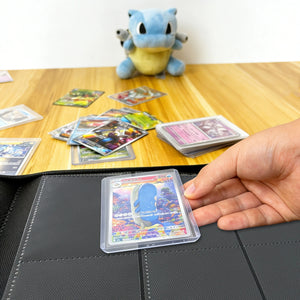 Hand placing a toploadered Pokemon card into a binder on a wood table beside a Blastoise plush and scattered cards