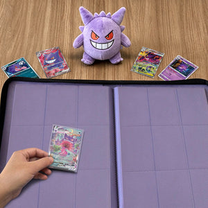 Hand placing holographic Pokemon card into 9-pocket binder beside Gengar plush on wood table